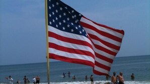 An American flag on a flagpole at a beach with people in the water and on the sand.