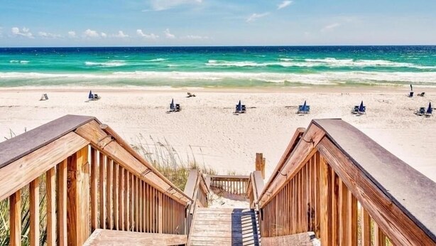 Eine Holztreppe führt zu einem Strand mit klarem türkisblauem Wasser und einem Sandufer.