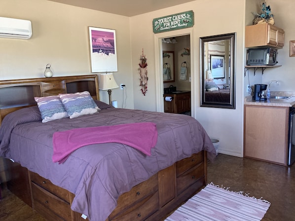 Un dormitorio con cama, mesita de noche, lámpara, letrero para cabañas turísticas, un baño visible a través de una puerta abierta y una zona de kitchenette.