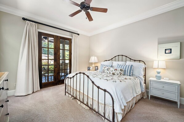 Une chambre à coucher comprenant un lit, une table de chevet, une lampe, un ventilateur de plafond et une vue sur l’extérieur, visible à travers une porte vitrée.