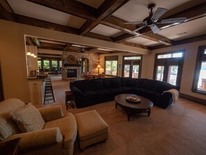 A spacious living room with a large sectional sofa, a coffee table, and a fireplace. There is a kitchen area in the background with a stone wall and a ceiling fan. - Gorgeous lakefront home with infinity pool, pickleball court, stunning lake view (Dawsonville)
