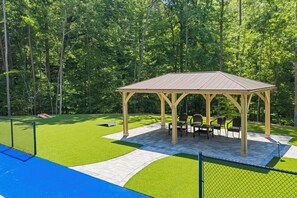 A gazebo with a tiled floor and outdoor furniture, surrounded by a fenced area with artificial grass and a playground. - Gorgeous lakefront home with infinity pool, pickleball court, stunning lake view (Dawsonville)
