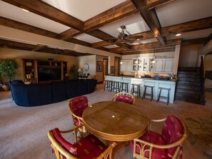 A spacious living room with a wooden ceiling, a large TV, and a dining area with red upholstered chairs. - Gorgeous lakefront home with infinity pool, pickleball court, stunning lake view (Dawsonville)
