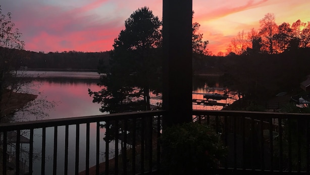 Vista del atardecer desde un balcón con vistas a un lago, árboles y barandilla de madera.