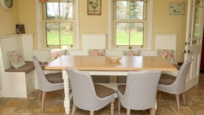 A dining room with a wooden table, wicker chairs, floral cushions, and a view of the garden through large windows. - Luxurious Cottage Set in Beautiful Irish Countryside. (Nenagh)