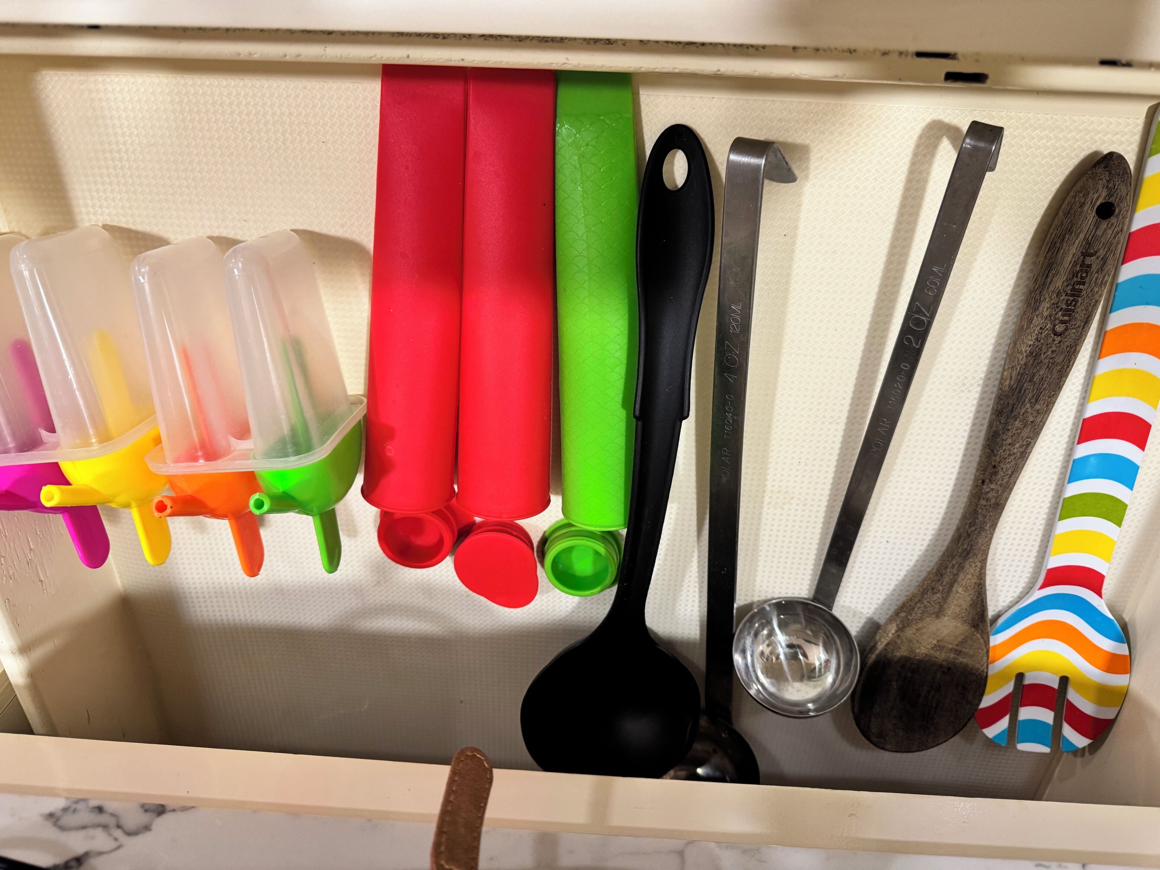 Un tiroir contenant divers ustensiles et récipients de cuisine, notamment des boîtes en plastique à couvercle, une louche noire, un verre doseur en métal et une cuillère en bois.