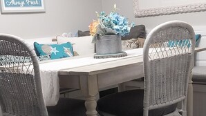 A dining area with a white table, wicker chairs, a potted plant, and wall decorations including a sign and a framed picture.