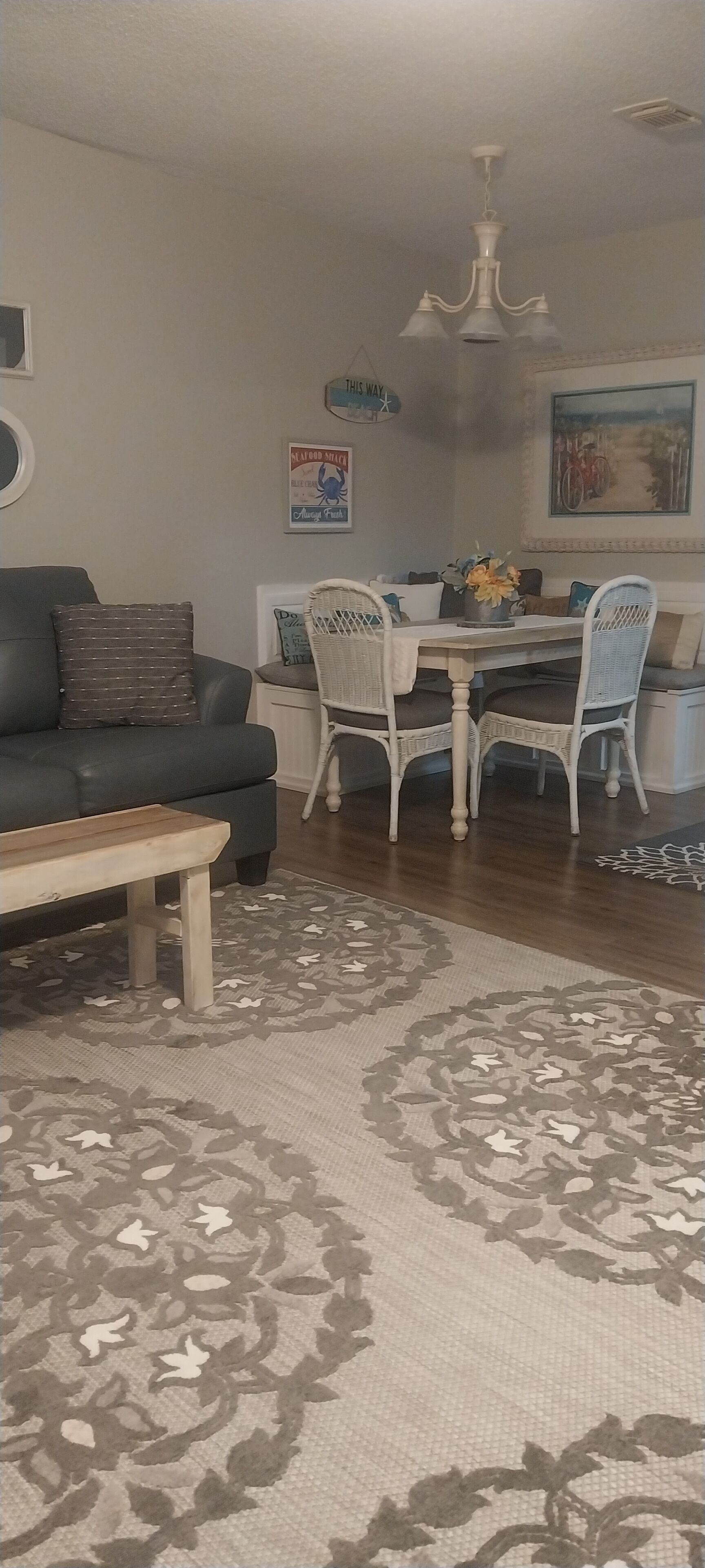 Una sala de estar con un sofá oscuro, una mesa de centro de madera, un tapete estampado, un comedor con sillas de mimbre blancas, una araña y un cuadro en la pared.