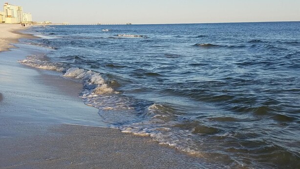 A beach with waves gently lapping onto the shore, a pier extending into the water, and buildings in the background.