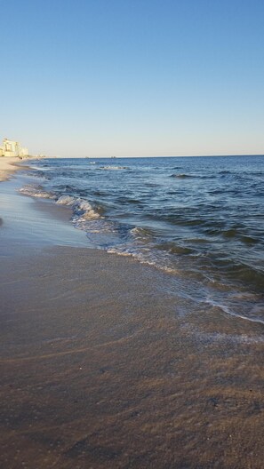 A beach with waves gently lapping onto the shore, a pier extending into the water, and buildings in the background.