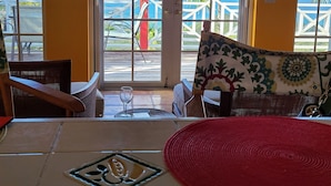 Una stanza con pavimento piastrellato, un divano con un cuscino rosso, un cuscino decorato e vista sull'oceano attraverso ampie finestre.