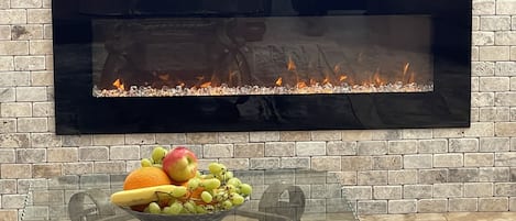 Un salon moderne avec une cheminée, une table en verre surmontée d’un panier de fruits et un mur en briques.