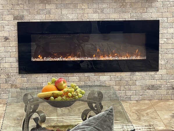 A modern living room with a fireplace, a glass table with a fruit bowl, and a brick wall.