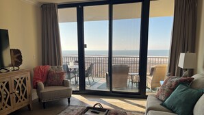 Un salon comprenant un canapé, un fauteuil et un pouf, une petite table avec un plateau, et une vue sur l’océan par une porte coulissante.