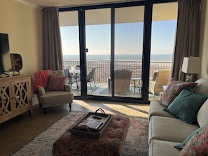 A living room with a sofa, armchair, and ottoman, a small table with a tray, and a view of the ocean through a sliding door.