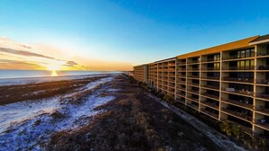Un hôtel au design moderne, doté de plusieurs étages et de balcons, situé à proximité d’une plage offrant une vue sur le coucher de soleil sur l’océan.