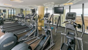 Une salle de sport  équipée de tapis de course, d’elliptiques et de vélos d’appartement, de grandes fenêtres offrant une vue sur la plage et d’un design moderne.