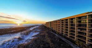A hotel with a modern design, featuring multiple floors and balconies, located adjacent to a beach with a view of the sunset over the ocean. - TOP FLOOR with PENTHOUSE views! BEACHFRONT- 2 BDRM-2 BATH, 2 POOLS and HOT TUB! (Dauphin Island)