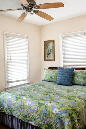 Un dormitorio con cama, ventilador de techo y dos ventanas con persianas.
