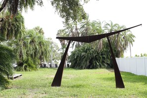 An outdoor sculpture with two dark metal supports and a flat top, surrounded by grass and palm trees.