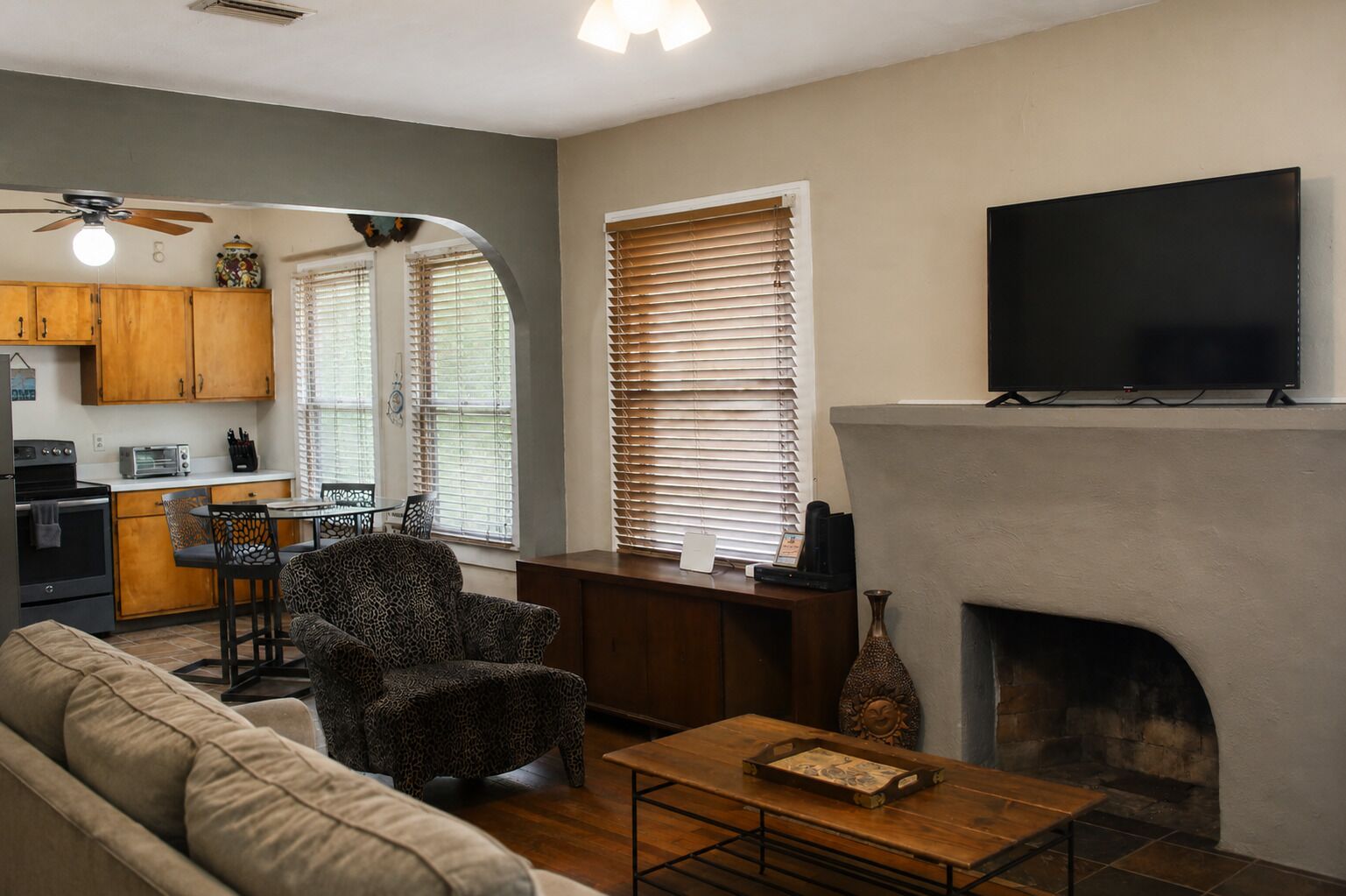 A living room with a fireplace, a television mounted above it, a kitchen area with wooden cabinets, and a dining table with chairs.