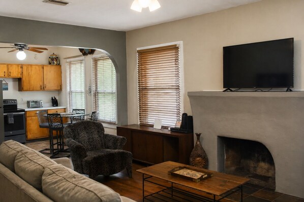 A living room with a fireplace, a television mounted above it, a kitchen area with wooden cabinets, and a dining table with chairs.