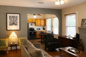 Una sala de estar con un sofá, un sillón estampado, una mesa de centro y una lámpara. Cuenta con una zona de cocina con armarios de madera, un refrigerador y un microondas.