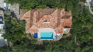 An aerial view of a house with a large swimming pool, surrounded by lush greenery.