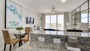 A modern kitchen with granite countertops, a dining area with wicker chairs, a large window with curtains, and a ceiling fan.