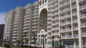 A multi-story hotel with balconies, a central arched entrance, and a row of palm trees in the foreground.