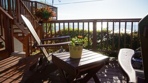 Une terrasse en bois avec une table et des chaises, un pot de fleurs et un escalier menant à un balcon d’où l’on peut admirer l’océan.