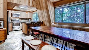 Interior de una cabaña de madera con cocina, comedor y grandes ventanales que dan a vistas arboladas.