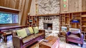 Interior acogedor de una cabaña con chimenea de piedra, estanterías de madera y un sofá de cuero.
