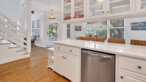 A kitchen with white cabinetry, a stainless steel dishwasher, and wicker chairs. - Summer Wind: Poolside Retreat Near the Beach & Village (St. Simons Island)