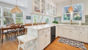 A modern kitchen with white cabinetry, a built-in dishwasher, and a dining area with wooden chairs and a white table. - Summer Wind: Poolside Retreat Near the Beach & Village (St. Simons Island)