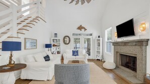 A spacious living room with a white staircase, a fireplace, and a television mounted above it. - Summer Wind: Poolside Retreat Near the Beach & Village (St. Simons Island)