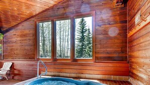 Interior de una cabaña de madera con jacuzzi, ventanas amplias y una silla.