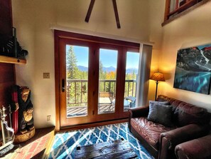 A cozy living room with a wooden door leading to a balcony, a brown leather sofa, a patterned rug, and a painting on the wall.