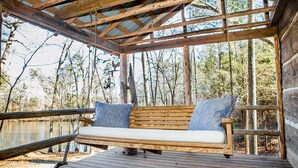 Una terraza de madera con un asiento tipo columpio y cojines, rodeada por una pérgola de madera y vistas a árboles y agua.