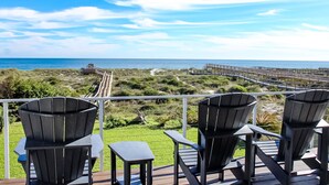 Eine Terrasse mit vier Adirondack-Stühlen und einem kleinen Tisch, mit Blick auf einen Strand mit einem hölzernen Steg und einer Promenade.