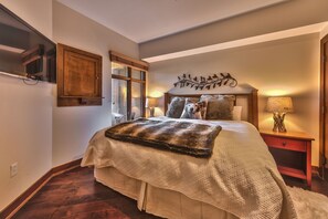 A bedroom with a large bed, a wooden headboard, a window, a nightstand with a lamp, and a wall-mounted shelf. - Perfect for Summer Balcony with Pool View Heart of Village (Park City)