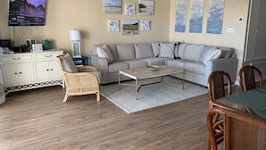 A living room with a grey sofa, wicker chair, wooden coffee table, and a dining table with chairs. - Beach Front Condo on the Strand, 101 Steps to the Beach  STR2021-00204 (Tybee Island)
