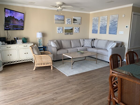 Een woonkamer met een grijze bank, een rieten stoel, een houten salontafel en een eettafel met stoelen.