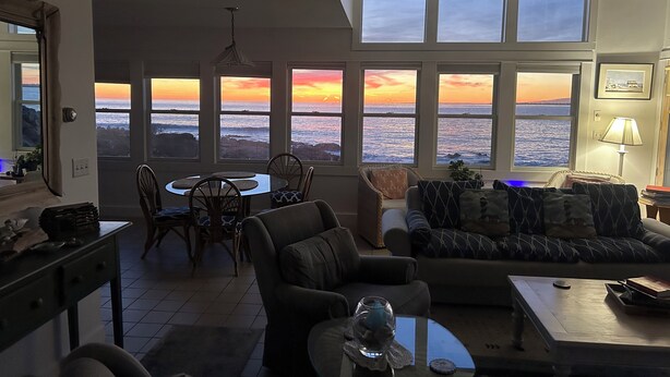 Un salon avec vue sur l’océan au coucher du soleil, comprenant un coin repas, des sièges et une lampe.