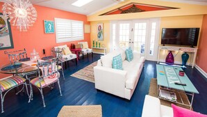 A living room with a white sofa, a glass dining table with chairs, a TV, and artwork on the walls. - A bit of Key West in New Port Richey: Bright Home w/ Pool & Boat Dock Access! (New Port Richey)