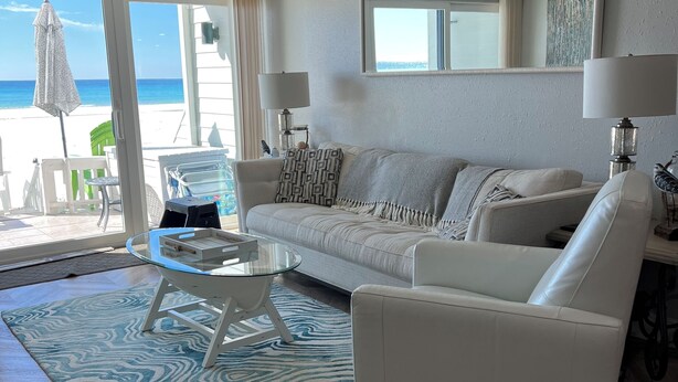 Una sala de estar con vista a la playa, un ventilador de techo, una mesa de centro de vidrio, un tapete estampado y un sofá blanco.