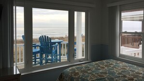 Une chambre avec une grande fenêtre donnant sur une plage, un ventilateur de plafond et un lit recouvert d’une courtepointe à motifs.