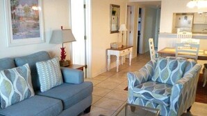 A living room with a sofa, armchair, and a glass coffee table. There is a painting on the wall and a lamp on a side table. - Beachfront - 2 bedrooms for price of one! (Pensacola)