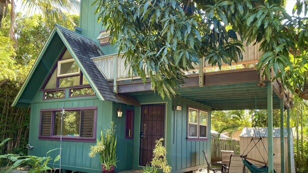 Ein zweistöckiges Haus mit grüner Fassade, einer Veranda mit Schaukelstuhl und einem kleinen Gartenbereich mit Blumentöpfen.