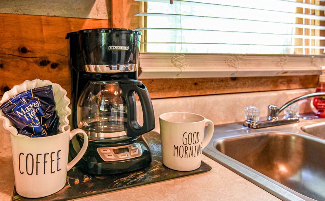 Een keuken met een koffiezetapparaat, een beker met de tekst 'COFFEE' en een andere met 'GOEDEMORGEN'.